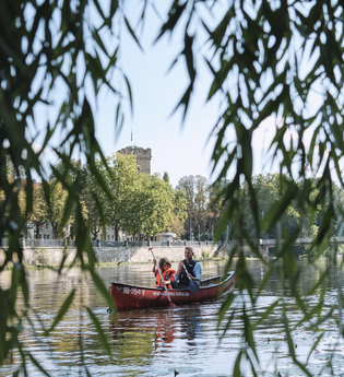 Das Bild wurde zwischen den hängenden Blättern einer Trauerweide aufgenommen. Zu erkennen sind zwei Personen in einem Kanu auf dem Fluss. Im Hintergrund erkennt man einen Turm aus Naturstein | © Heilbronn Marketing GmbH