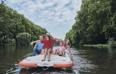 Im Fokus des Bildes ist ein weiß-orangenes Tretboot. An Bord befindet sich eine Familie mit zwei Kindern. Eines der Kinder sitzt ganz vorne auf dem Tretboot und breitet die Arme aus. Der Fluss ist an den Ufern mit Bäumen gesäumt | © Heilbronn Marketing GmbH