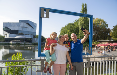 Eine Familie steht vor einem Geländer und fotografiert sich selbst durch einen blauen Bilderrahmen. Im Hintergrund sieht man die experimenta. Rechts im Bild sind die Neckarbühne mit roten Sonnenschirmen zu sehen | © Heilbronn Marketing GmbH