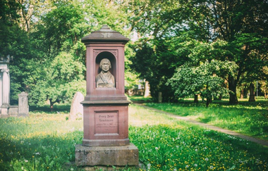 Im Vordergrund steht ein alter Grabstein aus rotem Stein. Im oberen Teil ist ein Abbild einer Person aus Stein und unten ein Schriftzug. Der Rasen unter dem Steingebilde ist schattig, dahinter ist ein sonniger Fleck. Daneben führt ein Weg durch den Wald.