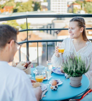 Das Bild zeigt zwei Personen, die ein Frühstück auf der Aussichtsfläche des Hafenmarktturms genießen. Es gibt Orangensaft, gekochte Eier und Brötchen. Im Hintergrund sieht man die Gebäude der Innenstadt. Die Atmosphäre ist fröhlich und locker.
