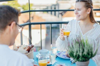 Das Bild zeigt zwei Personen, die ein Frühstück auf der Aussichtsfläche des Hafenmarktturms genießen. Es gibt Orangensaft, gekochte Eier und Brötchen. Im Hintergrund sieht man die Gebäude der Innenstadt. Die Atmosphäre ist fröhlich und locker.