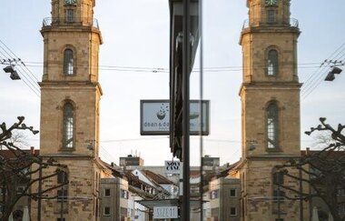 Das Bild zeigt den Hafenmarktturm, der sich in einer Glasfassade spiegelt. Der Himmel ist schön blau und leicht bewölkt, ein Mann geht auf der Straße entlang. Im Hintergrund sind Geschäfte, Straßenlampen und eine ruhige Stadtatmosphäre sichtbar.