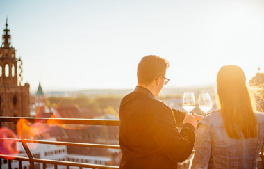 Das Bild zeigt zwei Personen am Geländer des Hafenmarktturms, die bei Sonnenuntergang mit Weingläsern anstoßen. Im Hintergrund sieht man die Stadt von der Aussichtsfläche des Hafemarktturms. Links sieht man die Kilianskirche