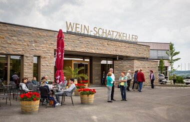 Einladende Szene vor einem Gebäude. Gäste sitzen entspannt an Tischen, umgeben von blühenden Blumenkübeln. Eine Gruppe steht am Eingang. Die Fassade ist aus Naturstein und auf dem Dach steht die Aufschrift "Wein-Schatzkeller" in goldenen Großbuchstaben.