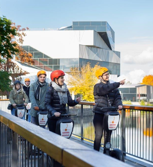 Das Bild zeigt eine Gruppe von Personen auf Segways. Sie fahren über eine Brücke. Die Stadtführerin zeigt auf etwas. Im Hintergrund sieht man ein Teil des Neckars und den Science Dome der Experimente.