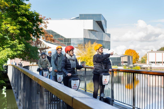Das Bild zeigt eine Gruppe von Personen auf Segways. Sie fahren über eine Brücke. Die Stadtführerin zeigt auf etwas. Im Hintergrund sieht man ein Teil des Neckars und den Science Dome der Experimente.