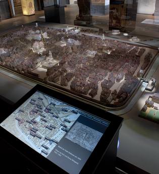 Das Bild zeigt ein detailliertes Modell der Stadt Heilbronn. Das Modell ist in einem Museum auf einem Tisch ausgestellt, neben einem interaktiven Touchscreen, der zusätzliche Informationen und Karten der Stadt anzeigt.