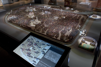 Das Bild zeigt ein detailliertes Modell der Stadt Heilbronn. Das Modell ist in einem Museum auf einem Tisch ausgestellt, neben einem interaktiven Touchscreen, der zusätzliche Informationen und Karten der Stadt anzeigt.