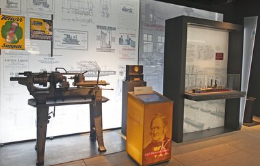 In der Ausstellung steht eine alte Drehmaschine. An der Wand sind historische Plakate und Skizzen. Neben der Drehmaschine befindet sich ein Ausstellungsstück mit einem Porträt von Carl Heinrich Knorr. Daneben ist eine Vitrine mit einem Schiffsmodell darin