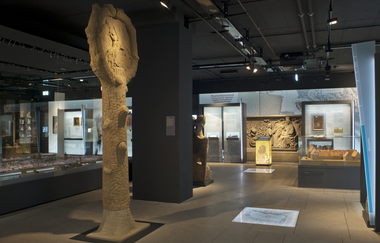 Das Bild zeigt den Innenraum eines Museums. Im Vordergrund steht eine steinerne Säule. Im Hintergrund sind Vitrinen mit kleineren Artefakten zu sehen. Der Raum ist modern gestaltet mit dunklen Wänden und indirekter Beleuchtung.