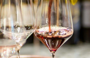 Das Bild zeigt drei Weingläser auf einem Holzfass. Aus einer dunklen Flasche wird Rotwein in eines der Gläser gegossen. Der Hintergrund ist verschwommen.