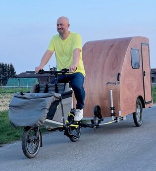 Ein Mann mit Jeans und gelbem T-Shirt bekleidet fährt auf einer asphaltierten Strasse mit einem Lastenfahrrad an dem ein kleiner Fahrradwohnwagen aus Holz angehängt ist | © Dennis Hirschmann