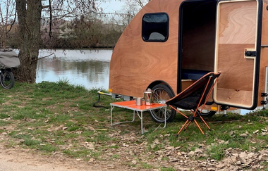 Fahrradwohnanhänger steht am Ufer eines Sees im Grünen mit einem kleinem roten Klapptisch und einem bequemen Klappstuhl davor | © Dennis Hirschmann
