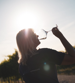 Ein Frau mit längeren, blonden Haaren die ein Glas Weißwein trinkt im Gegenlicht aufgenommen. Schöne Lichtstimmung im Sonnenuntergang mit ein Weinbergen im Hintergrund. | © Nicole Halter