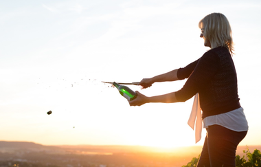 Nicole Halter sabriert eine Champagnerflasche bei Sonnenuntergang auf einem Weinberg. Der fliegende Korken und das spritzende Getränk betonen die Dynamik. Die warme Beleuchtung und die weite Landschaft schaffen eine feierliche, elegante Stimmung. | © DieWeinschmeckerin Nicole Halter