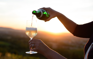 Eine Hand schenkt Weißwein aus einer grünen Weinflasche in ein Weinglas ein. Die Szene spielt bei Sonnenuntergang auf einem Weinberg. Im Hintergrund taucht der stimmungsvolle Untergang die Szene in ein warmes Licht. | © DieWeinschmeckerin Nicole Halter