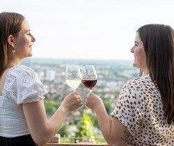 zwei junge Frauen stehen an einem Holzgeländer und stoßen mit Wein an. das Bild wurde auf einer Anhöhe aufgenommen und zeigt verschwommen die Weinreben und die Stadt im Hintergrund