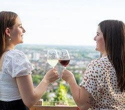 zwei junge Frauen stehen an einem Holzgeländer und stoßen mit Wein an. das Bild wurde auf einer Anhöhe aufgenommen und zeigt verschwommen die Weinreben und die Stadt im Hintergrund