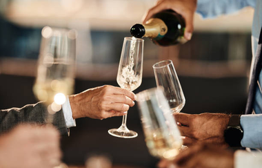 4 Männerhände halten Sektgläser in der Hand die teilweise befüllt sind, eines wird gerade mit einer Flasche befüllt