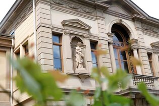 Das Bild zeigt einen Ausschnitt eines großen Gebäudes mit detailreichen Verzierungen an der Außenfassade. In der Mitte des Hauses ist ein Balkon mit großem Fenster und braunen Rahmen. Im Vordergrund sind dünne  Äste mit grünen Blättern zu sehen.
