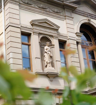 Das Bild zeigt einen Ausschnitt eines großen Gebäudes mit detailreichen Verzierungen an der Außenfassade. In der Mitte des Hauses ist ein Balkon mit großem Fenster und braunen Rahmen. Im Vordergrund sind dünne  Äste mit grünen Blättern zu sehen.