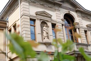 Das Bild zeigt einen Ausschnitt eines großen Gebäudes mit detailreichen Verzierungen an der Außenfassade. In der Mitte des Hauses ist ein Balkon mit großem Fenster und braunen Rahmen. Im Vordergrund sind dünne  Äste mit grünen Blättern zu sehen.