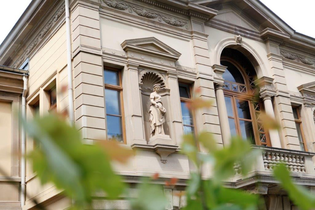 Das Bild zeigt einen Ausschnitt eines großen Gebäudes mit detailreichen Verzierungen an der Außenfassade. In der Mitte des Hauses ist ein Balkon mit großem Fenster und braunen Rahmen. Im Vordergrund sind dünne  Äste mit grünen Blättern zu sehen.
