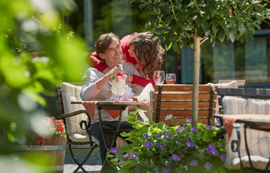 Auf dem Bild sitzt ein Mann mit Weinglas in der Hand auf einem Gartenstuhl aus Holz und Stahl. Eine Frau umarmt den Mann von hinten. Neben ihnen ist ein kleiner Baum und vor ihnen steht ein Blumenkübel mit lilanen Blüten. Das Bild wirkt friedlich und nett
