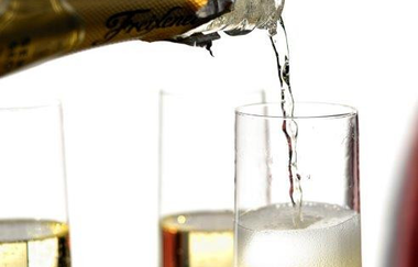 Das Bild zeigt einen Sektflaschenhals, aus dem Sekt in ein Sektglas fließt. In dem Glas  sprudelt die Kohlensäure. Dahinter stehen zwei weitere Gläser vor einem weißen Hintergrund.