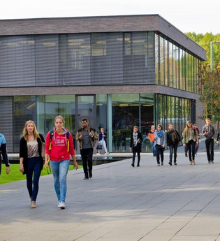 Im Hintergrund befindet sich ein zweistöckiges flaches Gebäude mit Rundumverglasung. Hinter dem Gebäude stehen große grüne Bäume. Auf dem gepflasterten Vorplatz sind verschieden große Gruppen Studenten unterwegs