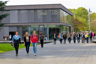 Im Hintergrund befindet sich ein zweistöckiges flaches Gebäude mit Rundumverglasung. Hinter dem Gebäude stehen große grüne Bäume. Auf dem gepflasterten Vorplatz sind verschieden große Gruppen Studenten unterwegs