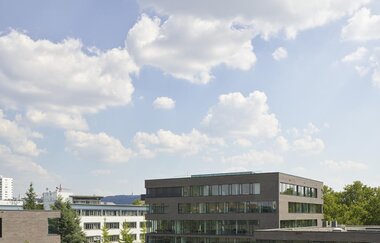 Im Fokus steht ein vierstöckiges braunes Gebäude mit Rundumverglasung. Rechts nach vorne versetzt steht dasselbe Gebäude etwas niedriger. Links davon befindet sich ein weißes, flaches, 3-stöckiges Gebäude . Auf dem Vorplatz sind Bäume gepflanzt