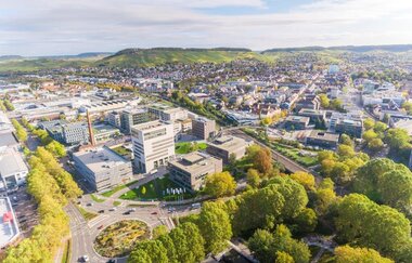 Im Fokus der Luftaufnahme liegt der Bildungscampus mit verschiedenen, unterschiedlich hohen Gebäuden. Links vor dem Bildungscampus befindet sich ein, in der Mitte bepflanzter, Kreisverkehr. Rechts im Bild sieht man unterschiedlich hohe grün belaubte Bäume