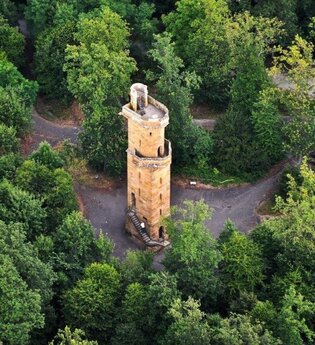 Im Fokus der Luftaufnahme steht ein runder Turm aus Naturstein auf asphaltiertem Grund umgeben von dichtstehenden, grünen Bäumen