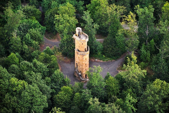 Im Fokus der Luftaufnahme steht ein runder Turm aus Naturstein auf asphaltiertem Grund umgeben von dichtstehenden, grünen Bäumen