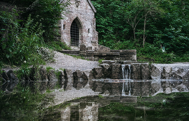 Inmitten des Waldes spiegelt sich ein kleines verwittertes Natursteinhaus mit bogenförmigem, vergittertem Eingang. Auf dem Dach befindet sich ein Steinkreuz. Das Ufer ist mit  verwitterten Steinen befestigt .