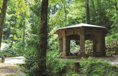 Inmitten des Waldes befindet sich ein Pavillon aus Naturstein mit flachem Metalldach. Im Vordergrund ist die bogenförmige Öffnung zu sehen durch die der Köpferbach kurzfristig unter dem Weg durchgeleitet wird.