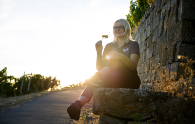 Nicole Halter sitzt entspannt auf einer Steinmauer am Rand eines Weinbergs und hält ein Glas Wein. Im Hintergrund scheint die Sonne warm durch das Grün, die Szene wirkt friedlich und idyllisch. Der Weg neben ihr führt leicht bergauf. | © Die Weinschmeckerin Nicole Halter