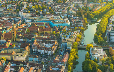 Das Bild zeigt die Luftaufnahme von einem Teil von Heilbronn. Rechts im Bild sieht man den Neckar gesäumt von Bäumen. Links ist ein Teil der Stadt rund um die Kilianskirche erkennbar | © Heilbronn Marketing GmbH/Roland Schweizer