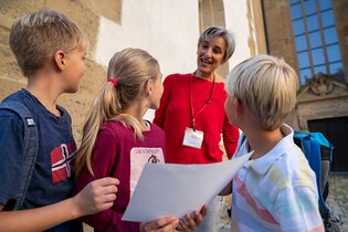 Das Bild zeigt eine Dame in rotem Langarm-Shirt. Drei Kinder stehen im Vordergrund, drehen sich nach hinten und schauen zu ihr auf. Im Hintergund ust eun großes Gebäude mit weißer Fassade und braunen Stein-Elementen zu sehen.