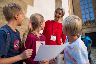 Das Bild zeigt eine Dame in rotem Langarm-Shirt. Drei Kinder stehen im Vordergrund, drehen sich nach hinten und schauen zu ihr auf. Im Hintergund ust eun großes Gebäude mit weißer Fassade und braunen Stein-Elementen zu sehen.