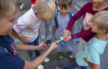 Das Bild fotografiert Kinder, die in einem Kreis stehen von oben. Die Kinder halten etwas in der Hand. Die Stimmung wirkt locker. Die Kinder schauen gespannt auf den Gegenstand.
