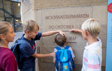 Das Bild zeigt vier Kinder vor einer Steinhausmauer. In der Mauer sind Hochwassermarken eingraviert. Zwei größere Kinder messen die Größe des Kleinsten in der Gruppe an einer Hochwassermarke. Die Kinder wirken interessiert und haben Spaß.