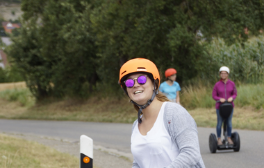 Lächelnde Frau fährt auf einem Segway auf der Straße. Im Hintergrund zwei weitere Personen auf einem Segway. | © GötzMotion