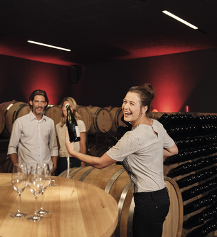 Das Bild zeigt einen dunklen Raum mit weinroten Wänden, vielen Holzfässern und Holzregalen für Flaschen. Im Vordergrund steht eine lachende Frau im Lichtpegel mit einer Flasche in der Hand. Neben ihr ist ein Tisch aus Holz mit leeren Sektgläsern.