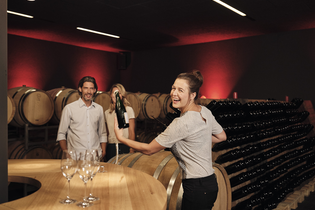 Das Bild zeigt einen dunklen Raum mit weinroten Wänden, vielen Holzfässern und Holzregalen für Flaschen. Im Vordergrund steht eine lachende Frau im Lichtpegel mit einer Flasche in der Hand. Neben ihr ist ein Tisch aus Holz mit leeren Sektgläsern.