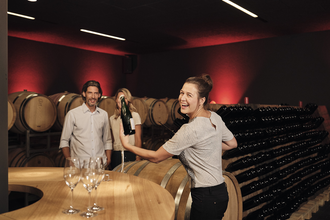 Das Bild zeigt einen dunklen Raum mit weinroten Wänden, vielen Holzfässern und Holzregalen für Flaschen. Im Vordergrund steht eine lachende Frau im Lichtpegel mit einer Flasche in der Hand. Neben ihr ist ein Tisch aus Holz mit leeren Sektgläsern.