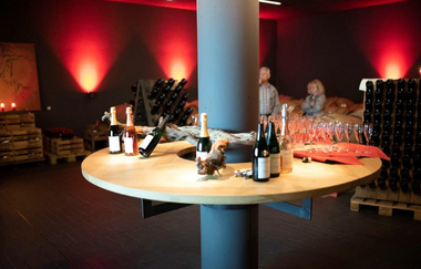 Es ist ein Raum mit gedämpfter Beleuchtung und dunkelroten Wänden. In der Mitte steht ein runder Tisch mit Sektflaschen, Gläsern und Snacks. Im Hintergrund sind Weinregale zu sehen, während zwei Personen die gemütliche Atmosphäre genießen.