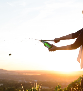 Nicole Halter sabriert eine Champagnerflasche bei Sonnenuntergang auf einem Weinberg. Der fliegende Korken und das spritzende Getränk betonen die Dynamik. Die warme Beleuchtung und die weite Landschaft schaffen eine feierliche, elegante Stimmung. | © DieWeinschmeckerin Nicole Halter